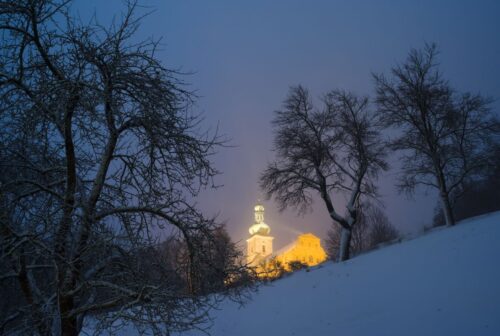 Verschneiter Hügel mit beleuchteter Kirche im Hintergrund, nächtliche Aufnahme.