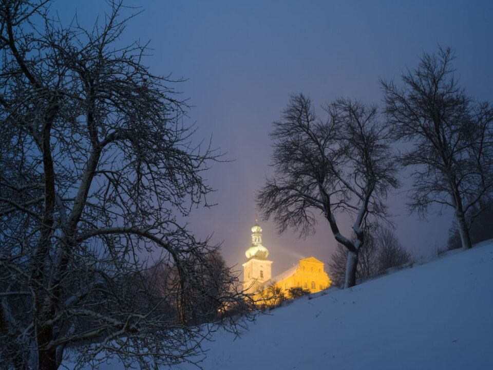 Verschneiter Hügel mit beleuchteter Kirche im Hintergrund, nächtliche Aufnahme.