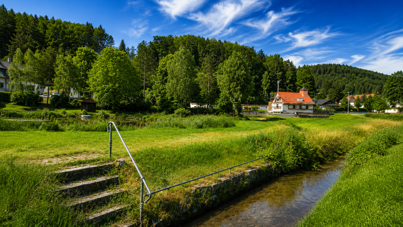 Natürliches Kneippbecken im Back mit steinernen Stufen und Handlauf, im Hintergrund Häuser des Dorfes, viel grüne Wiese und viele Bäume.