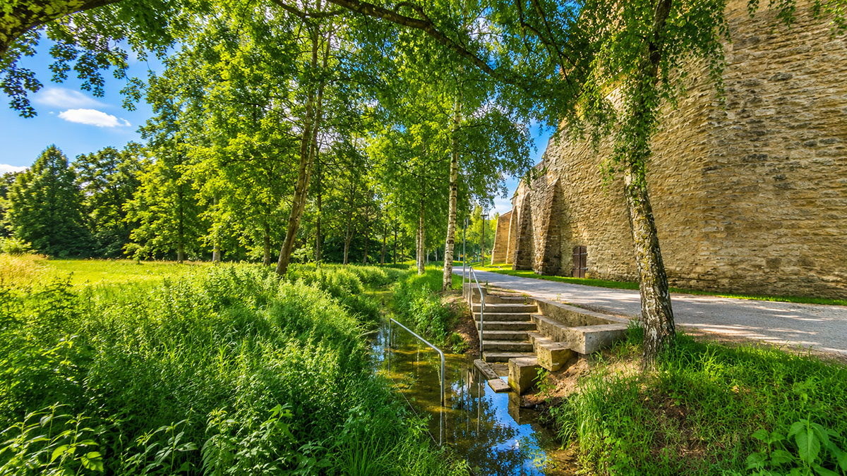Natürliches Kneippbecken im Bach mit Steinstufen und Handläufen inmitten einer Wiese. Daneben eine kleine Straße und die Mauern einer Burg.