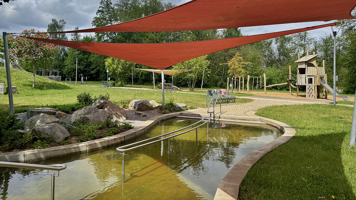 Natürliches Kneippbecken mit roten Sonnensegeln darüber auf einem Kinderspielplatz, im Hintergrund ein Turm mit Rutsche.